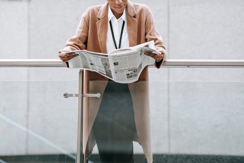 Free Crop content African American businesswoman in formal clothes reading fresh newspaper and leaning on glass railing of contemporary office building Stock Photo