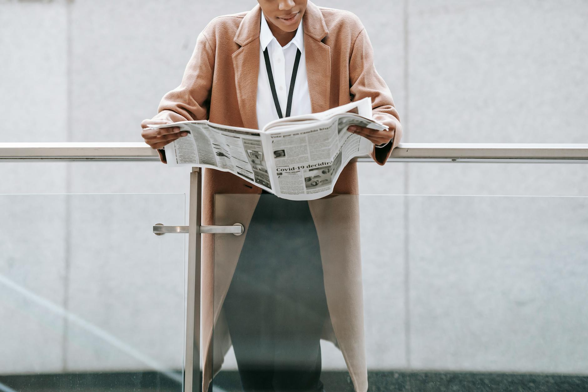 Crop content African American businesswoman in formal clothes reading fresh newspaper and leaning on glass railing of contemporary office building