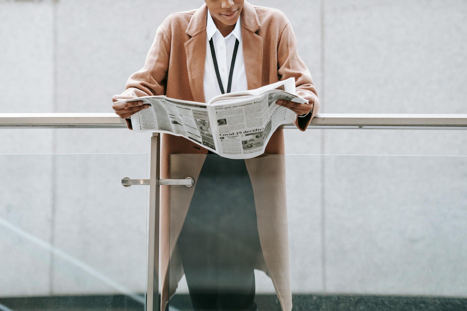 Crop content African American businesswoman in formal clothes reading fresh newspaper and leaning on glass railing of contemporary office building