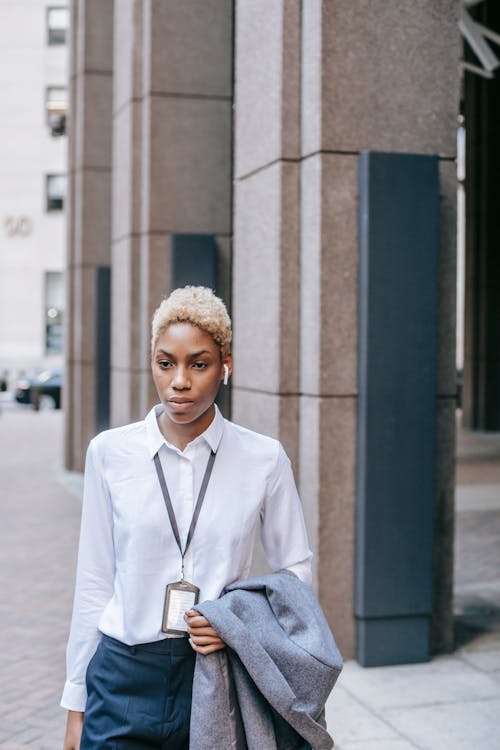 Free Stylish confident African American businesswoman in formal wear and earbuds walking on modern city street and carrying jacket on hand Stock Photo