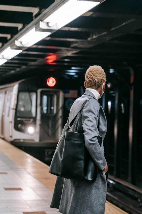 Back view anonymous female in gray coat and mask standing on metro station platform near arriving train