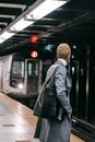 Back view anonymous female in gray coat and mask standing on metro station platform near arriving train