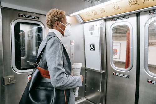 Back view contemplative young African American female in stylish wear and face mask standing with handbag and newspaper and riding subway train
