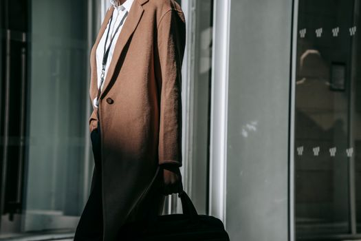 Side view crop anonymous businesswoman in formal clothes and coat carrying briefcase and walking along modern glass building
