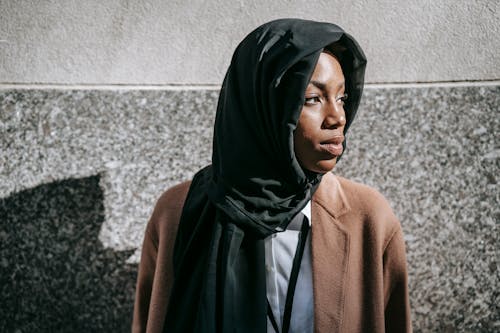 Mujer En Hijab Negro Y Abrigo Marrón De Pie Junto A La Pared Blanca