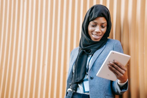 Cheerful young African American female in traditional Islamic hijab and formal wear browsing modern tablet while leaning on brown wall