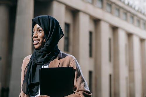 Cheerful Muslim black woman with laptop standing outside modern building