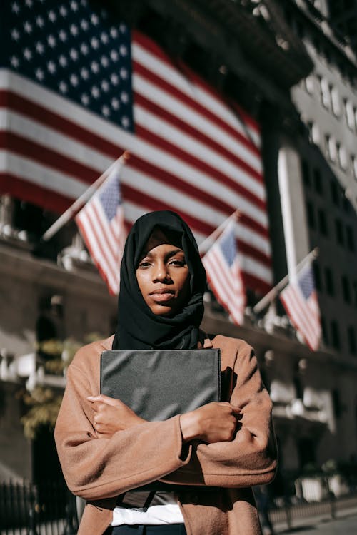 Serious young African American female in traditional Islamic headscarf holding black folder and looking at camera calmly while standing outside contemporary building decorated with US national flags