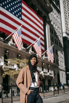 Cheerful Muslim female ambassador wearing traditional headscarf looking away while standing with hand in pocket near building with American flags