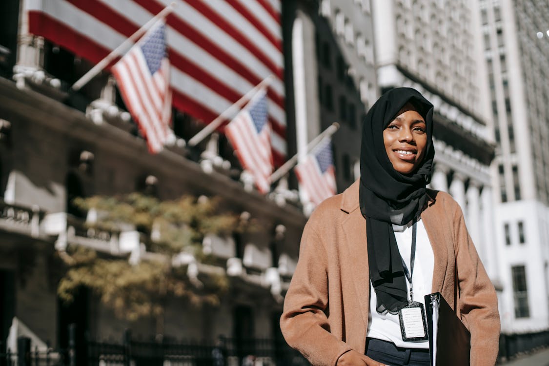 Free From below of cheerful African American female ambassador with folder wearing hijab and id card looking away while standing near building with American flags on blurred background Stock Photo