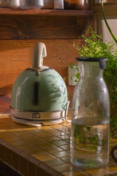 A retro green kettle and glass water jug on a sunlit kitchen counter with herbs.