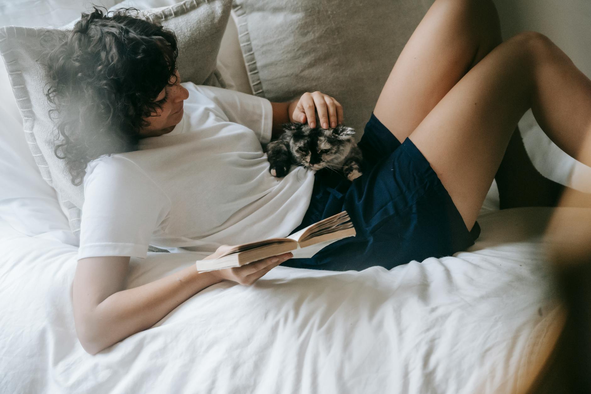 Side view content young female in casual clothes stroking cute cat while resting on cozy bed with book