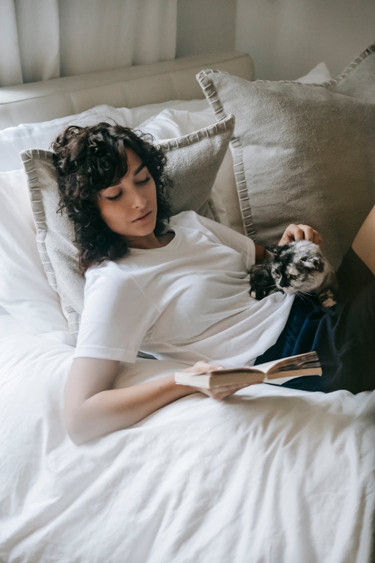 Calm Woman Reading Book And Caressing Cat On Bed