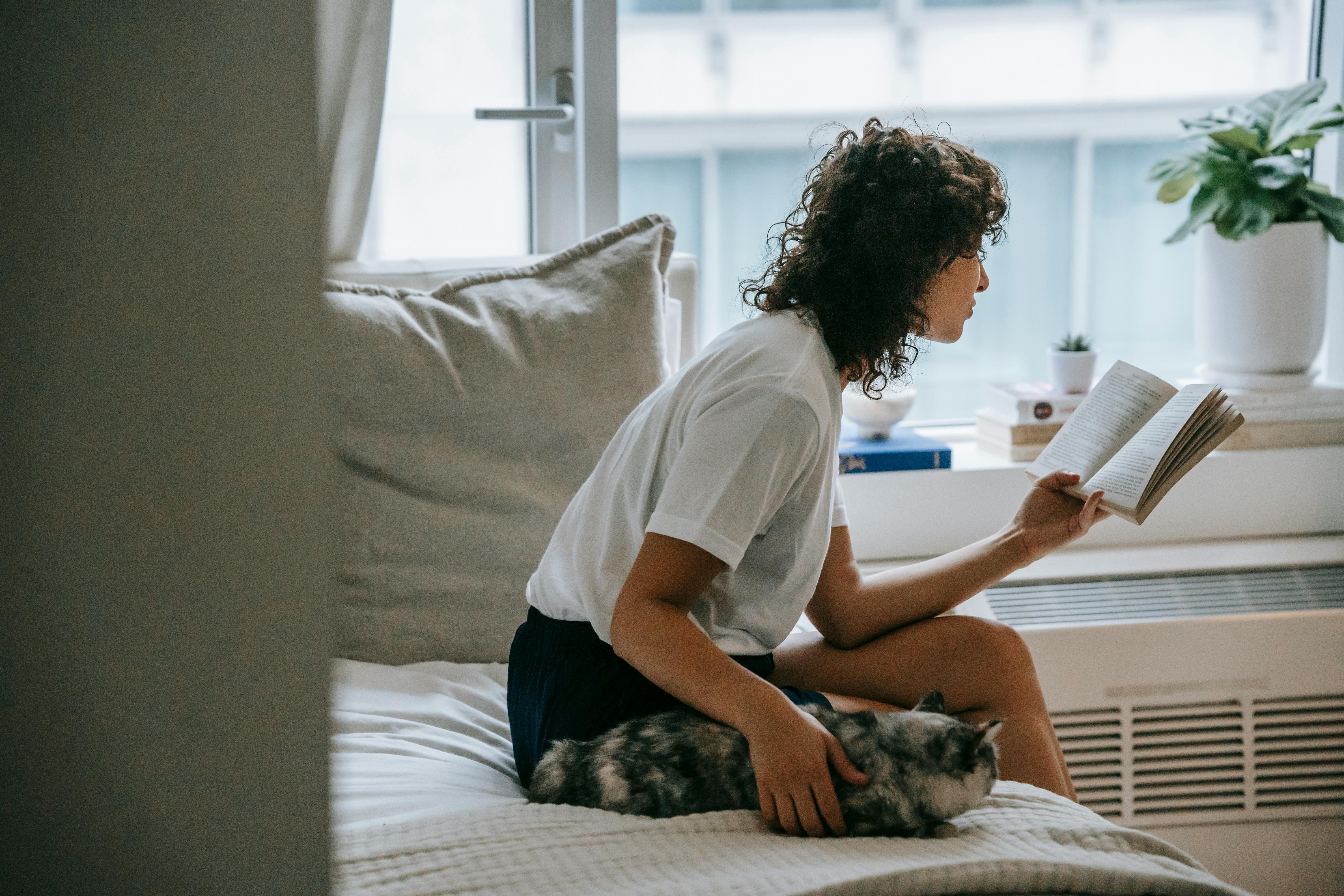 focused woman reading book on bed near cat