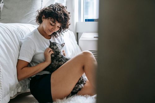 Free Side view cheerful pretty brunette with curly hair cuddling adorable hairy cat while sitting on floor against soft bed in light bedroom Stock Photo