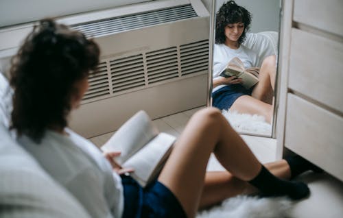 Mirror reflection of calm brunette reading book on floor in cozy light bedroom at home