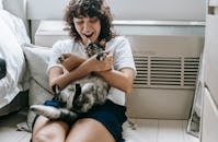 Happy female with dark hair caressing adorable dappled cat while sitting on floor with hands crossed near radiator and bed in room at home