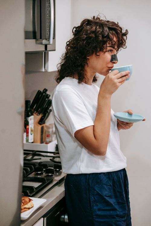 Free Young curly female with cosmetic nose strip enjoying cup of coffee leaning on kitchen counter Stock Photo