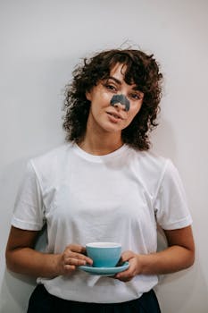 Charming female with curly hair treating skin with blackhead nose mask while standing with cup of hot drink