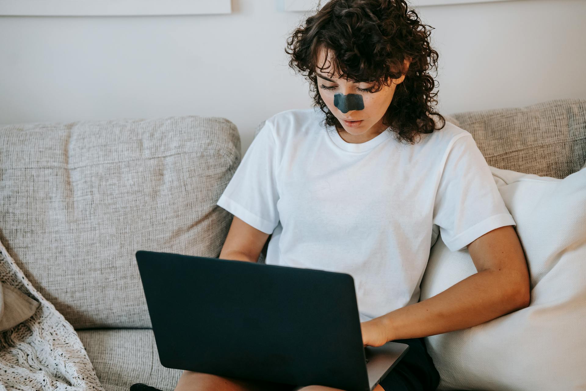 Focused woman with charcoal nose mask typing on netbook on sofa