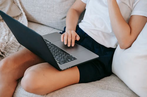 Free From above of crop unrecognizable self employed young lady in casual clothes sitting on comfortable couch and working online on laptop at home Stock Photo