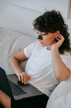 Young woman with nose patch working on laptop at home, combining work with self-care.
