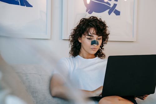 Focused young female freelancer with curly hair in casual outfit sitting on sofa with anti acne patch on nose and working remotely on laptop