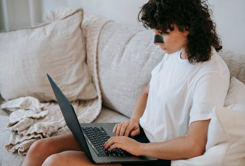 Side view of crop serious young self employed woman in casual clothes sitting on couch and typing on laptop during daily skin care routine at home