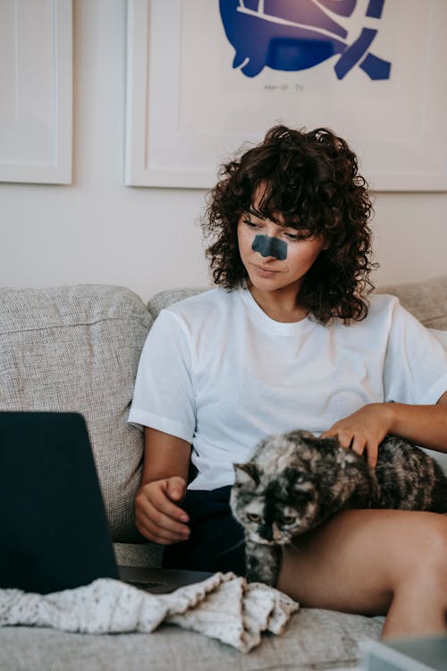 Calm young lady in casual clothes with dark curly hair and charcoal anti acne mask on nose watching  interesting movie on laptop and stroking cat while relaxing on sofa