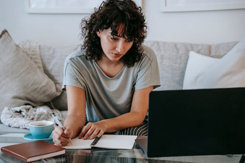 Serious young lady writing in notebook during online studies on laptop at home