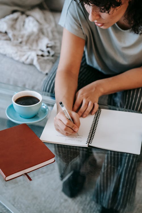 Free From above of crop young female student in casual clothes taking notes in copybook and drinking coffee while sitting on couch and doing homework assignment Stock Photo