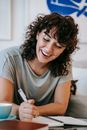 Cheerful young female student with curly dark hair in casual clothes sitting near cute cat and taking notes while doing homework assignment
