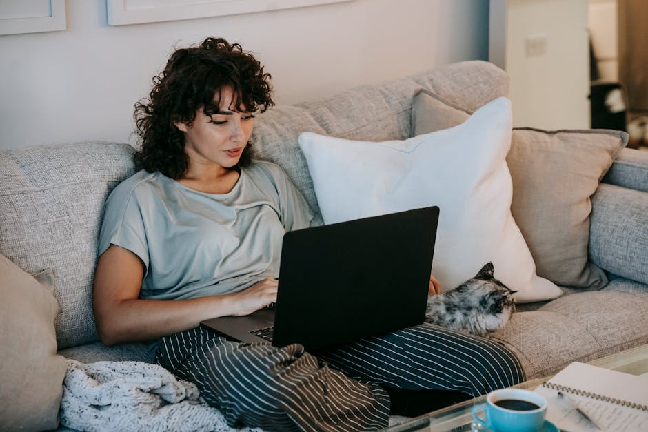 Clumping vs Crystal Litter: Odor Control Showdown A young woman with curly hair relaxes on a cozy sofa, working on her laptop with a cat beside her.