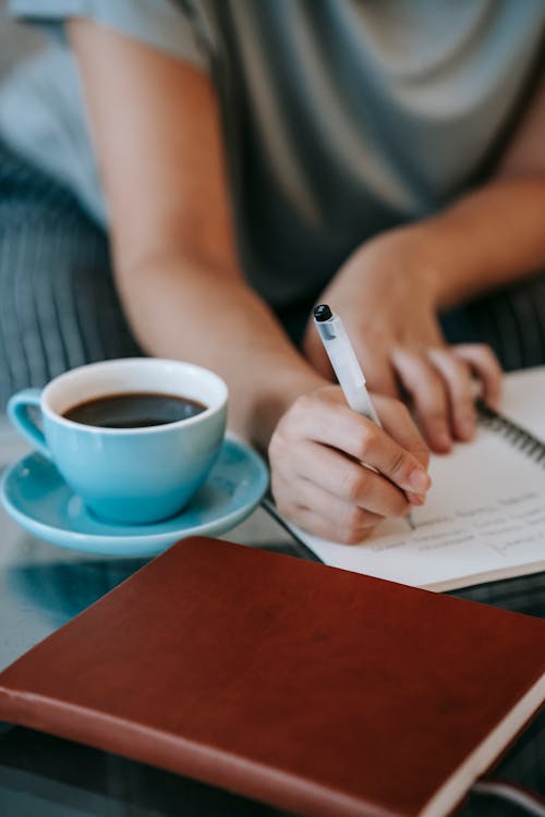 High angle of crop unrecognizable female author taking notes in diary and drinking cup of coffee while working remotely at home