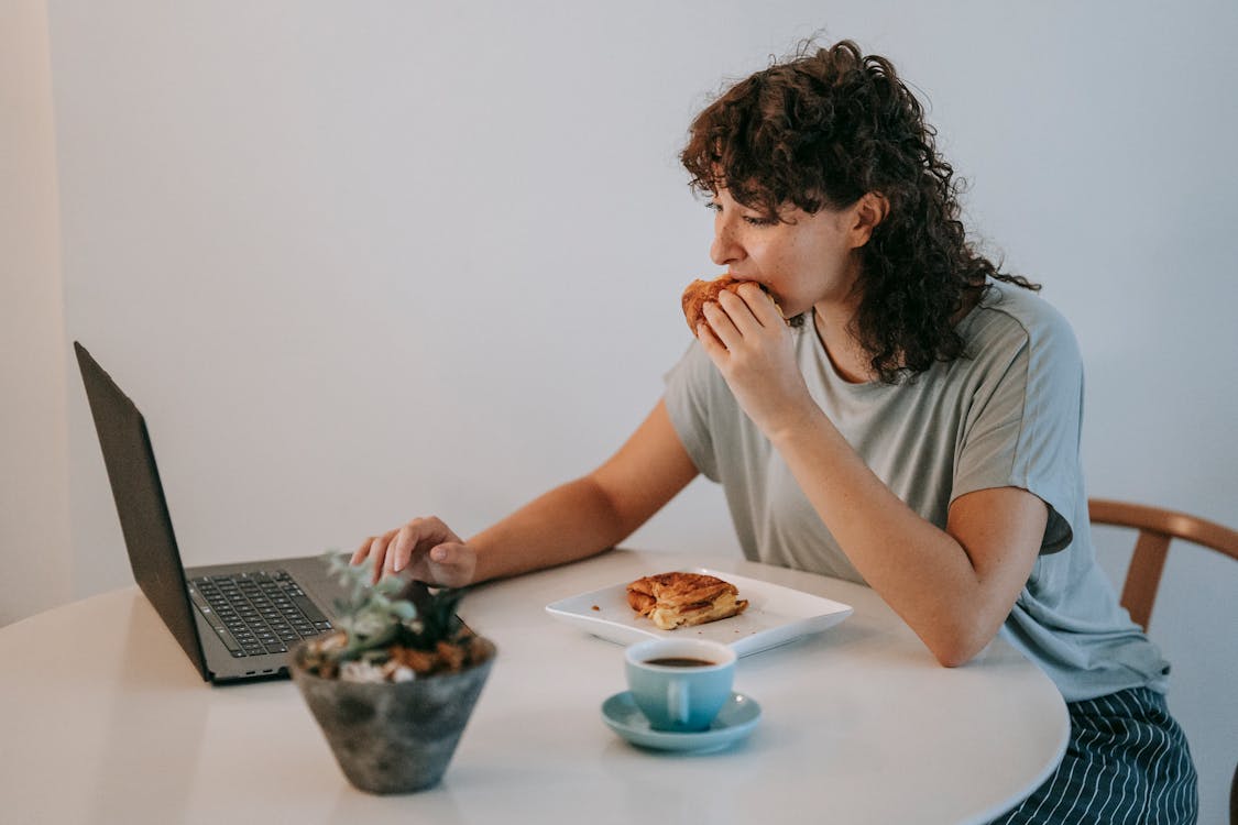 Free Young female with curly dark hair working with netbook while looking at screen and eating toasts with cup of coffee Stock Photo