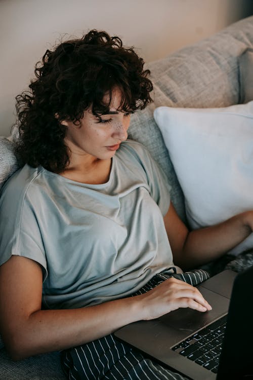 Free From above of concentrated young ethnic female freelancer with curly hair in casual clothes sitting on comfortable sofa and working remotely on laptop at home Stock Photo