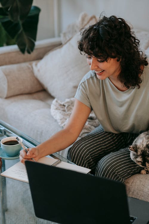 Cheerful young ethnic woman taking notes while doing homework using laptop