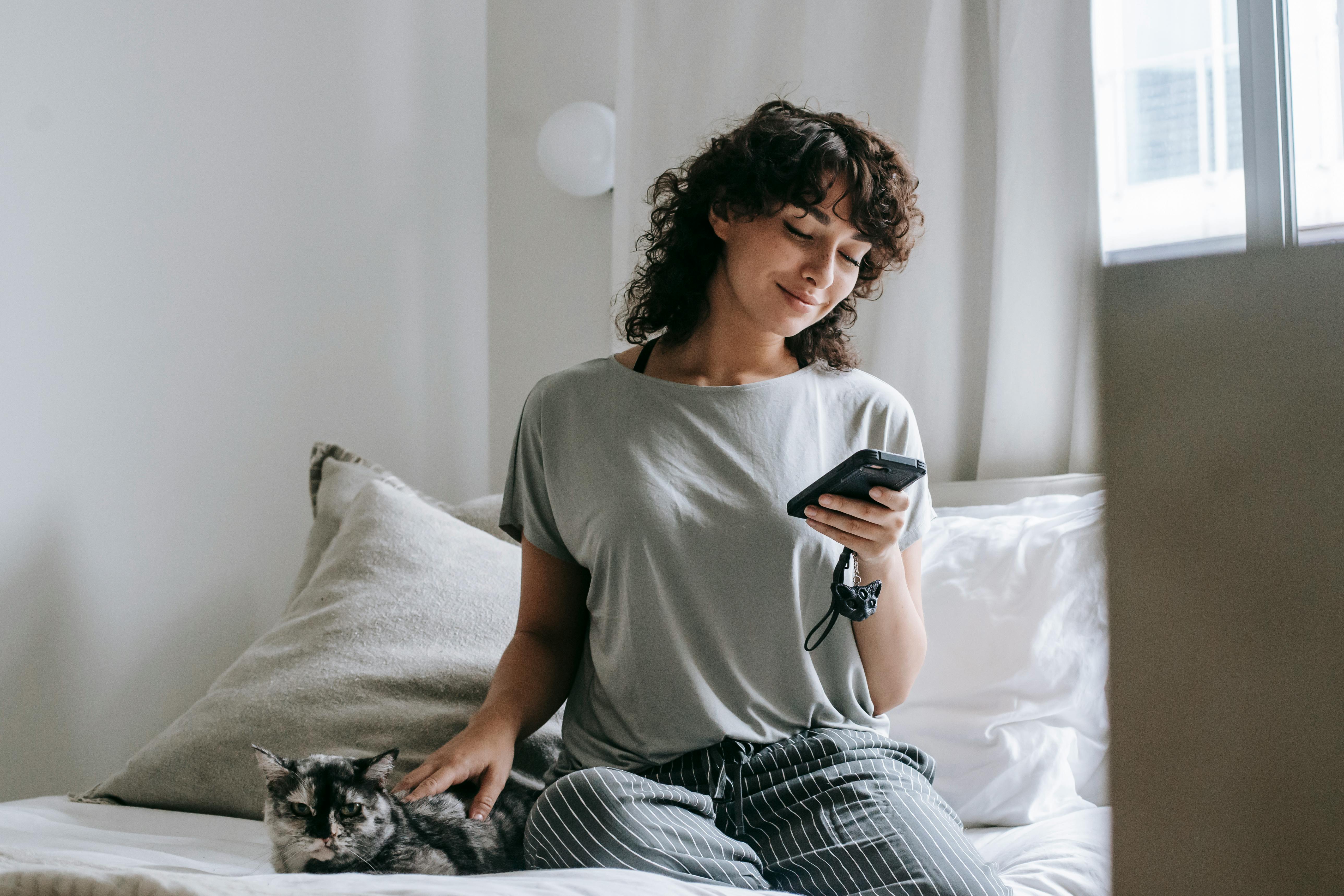 smiling young ethnic female messaging on smartphone and petting cat on bed