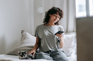 Free Smiling young ethnic female messaging on smartphone and petting cat on bed Stock Photo