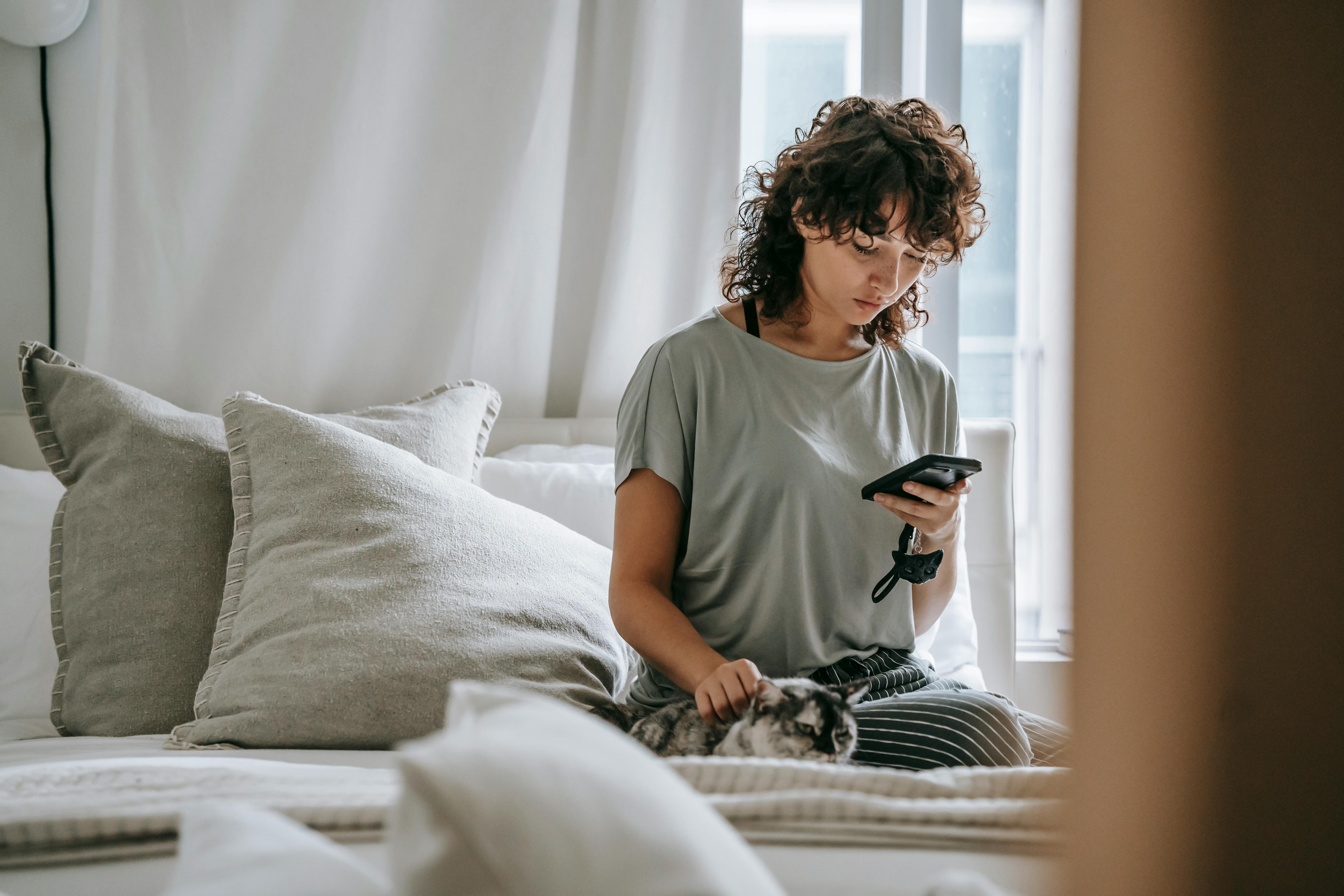 serious young ethnic lady reading message and petting cat on bed