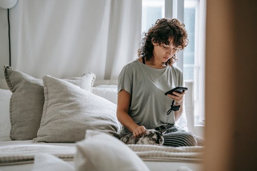 Concentrated young ethnic woman with curly hair in casual clothes messaging on mobile phone and stroking cat while relaxing on comfortable bed during weekend