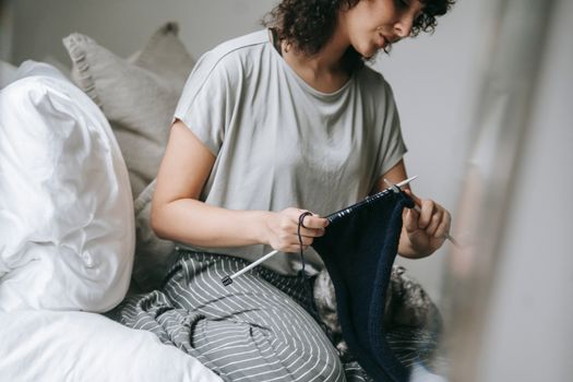 Crop young ethnic lady with curly hair in comfy clothes sitting on soft bed with cat and knitting with needles and yarn