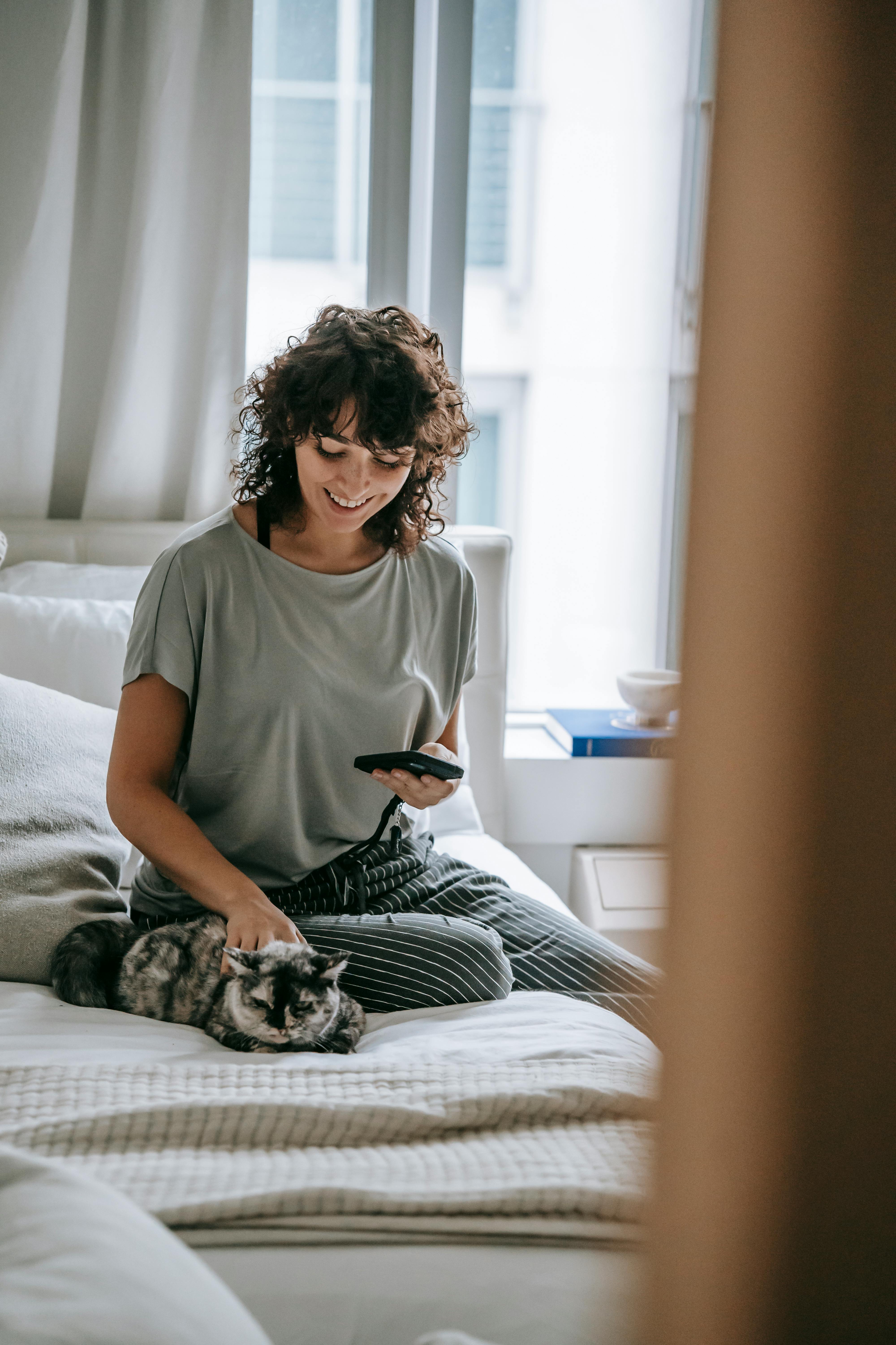 cheerful young female owner petting cat and using smartphone on bed