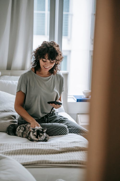 Cheerful young female owner petting cat and using smartphone on bed