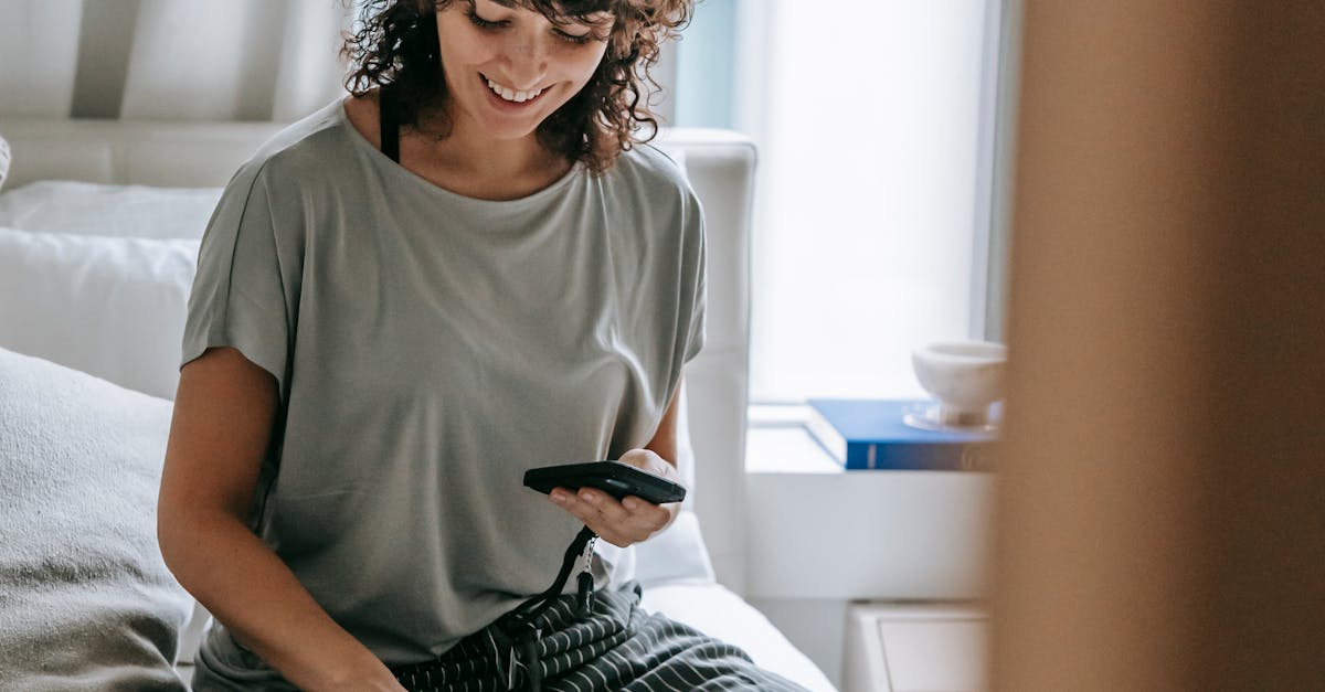 Cheerful young female owner petting cat and using smartphone on bed