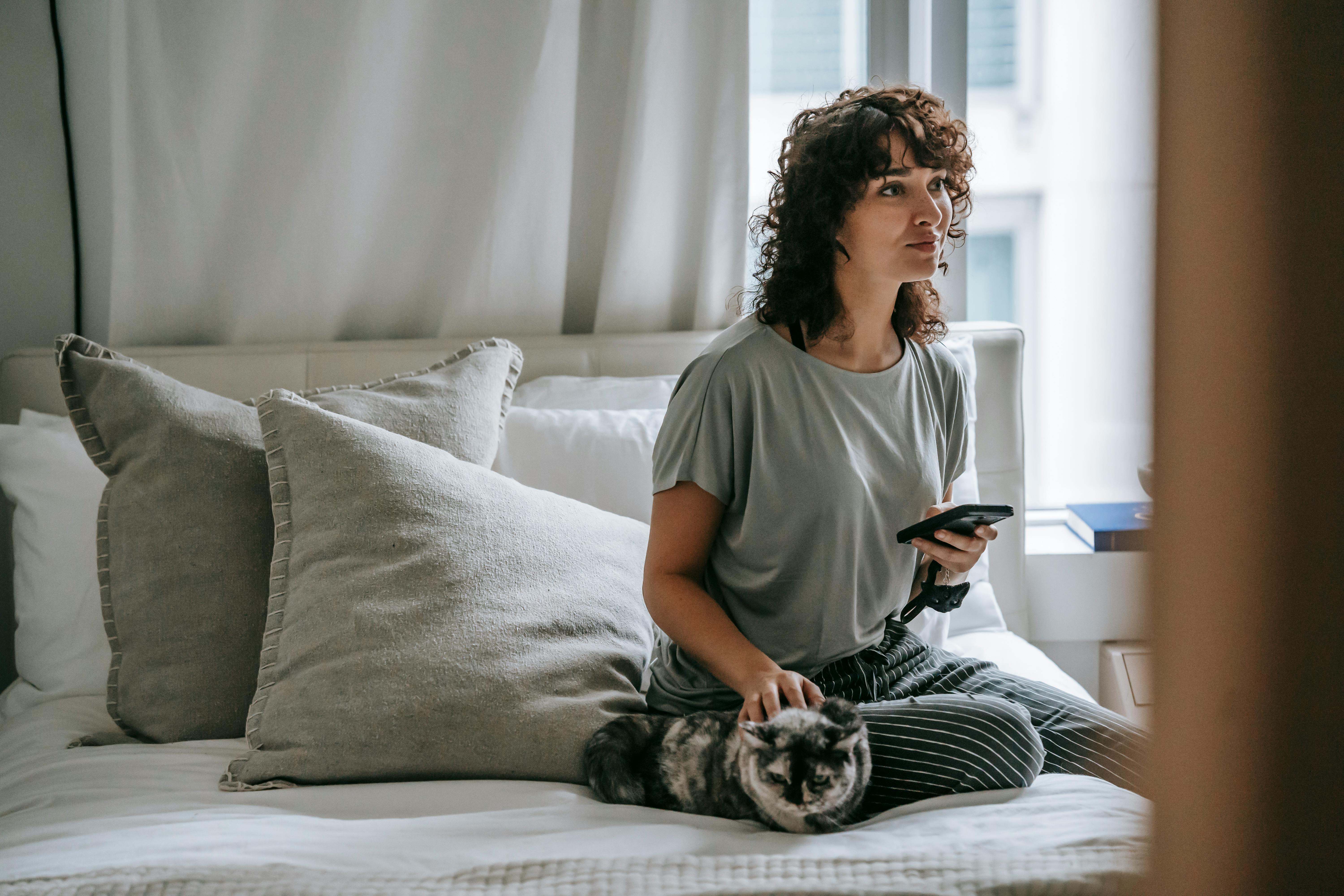 pensive young ethnic lady messaging on smartphone while relaxing on bed with cat