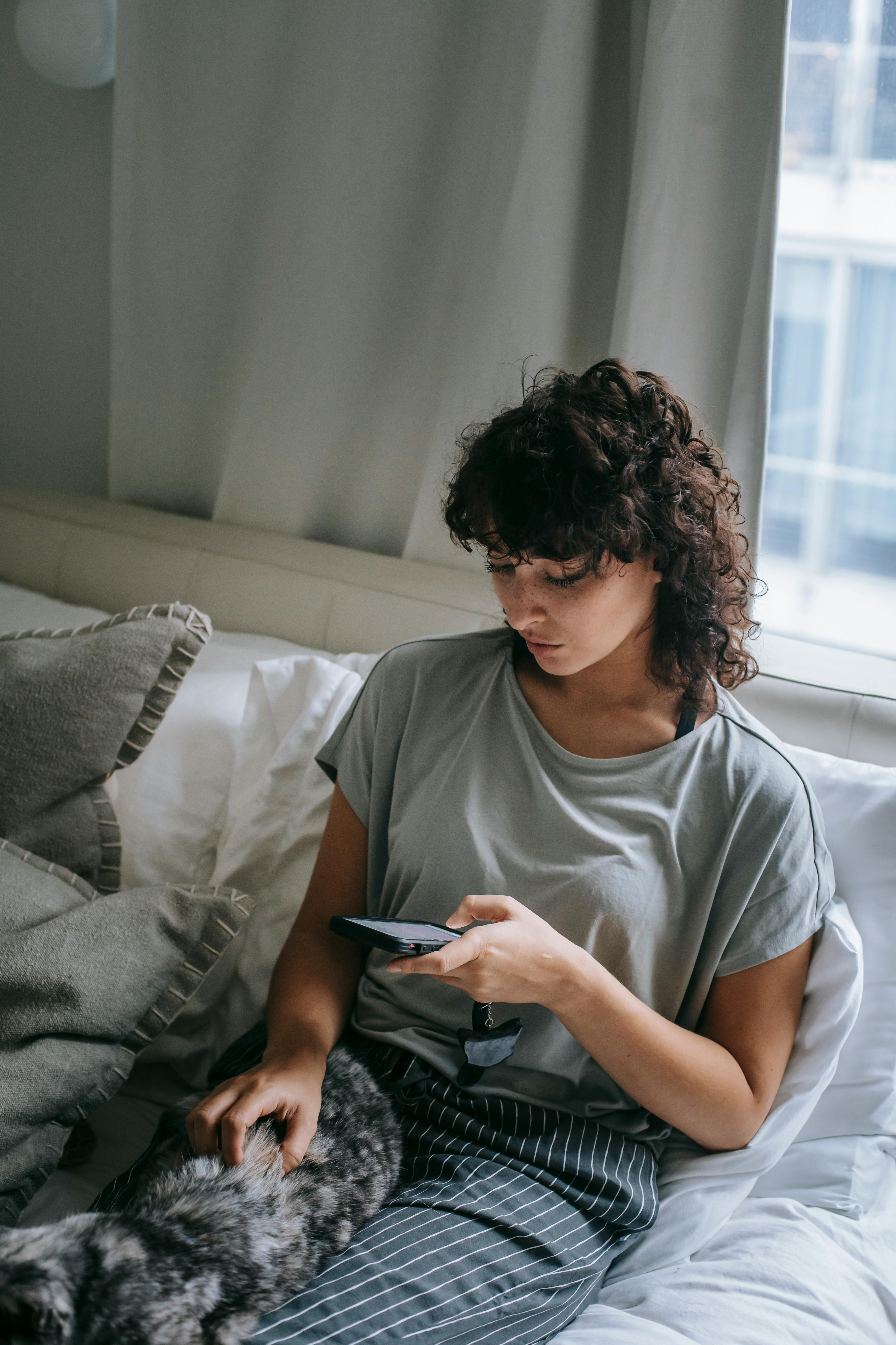 woman chatting on smartphone while caressing cat on bed