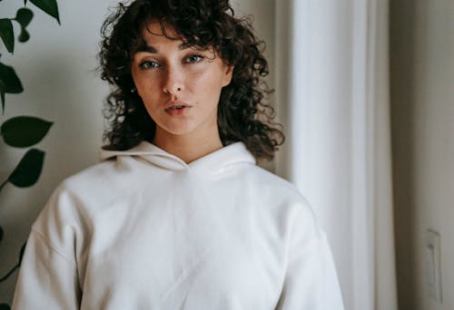 Crop adult female in soft wear with curly hair looking at camera near plant in house