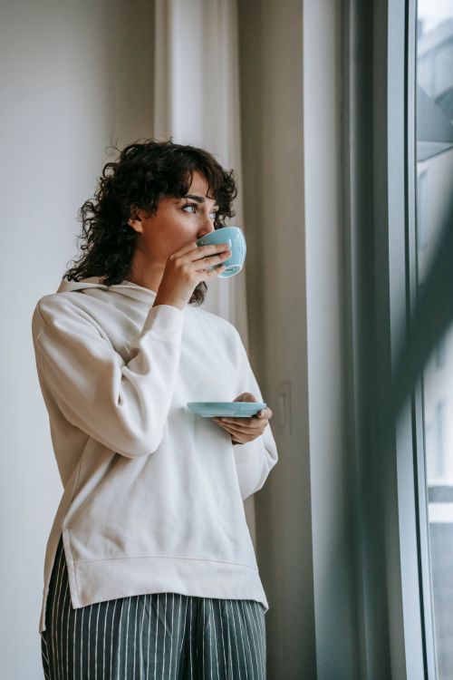 Free Low angle of female in casual wear drinking hot coffee in morning standing near panoramic window Stock Photo