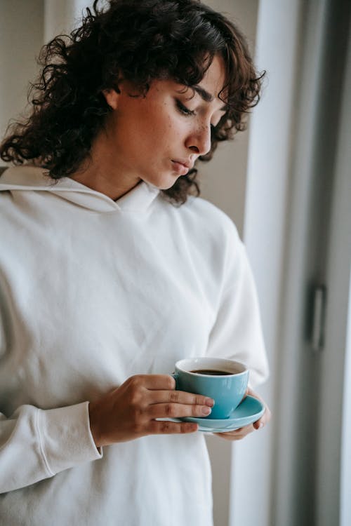 Disappointed woman with curly hair standing with ceramic mug of hot drink and looking down
